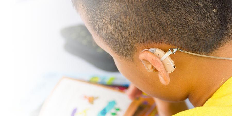 A close up focusing on the back of a child’s head and their hearing aid device in their ear. On the table in front of the child lays a paper with the child’s drawing of animals. The child is holding a crayon.